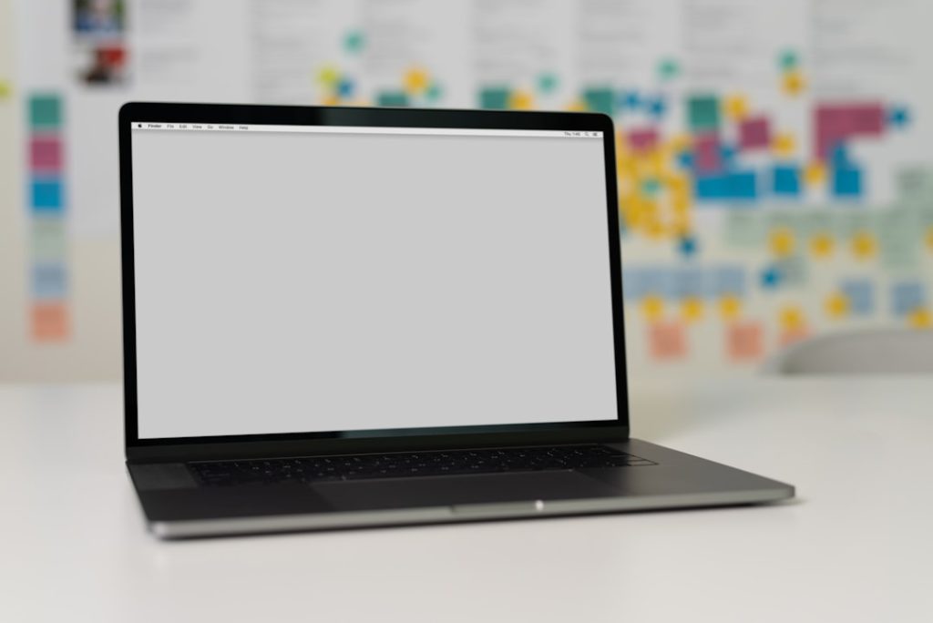 A laptop on a desk with an empty screen, background features sticky notes and papers organized on a wall.