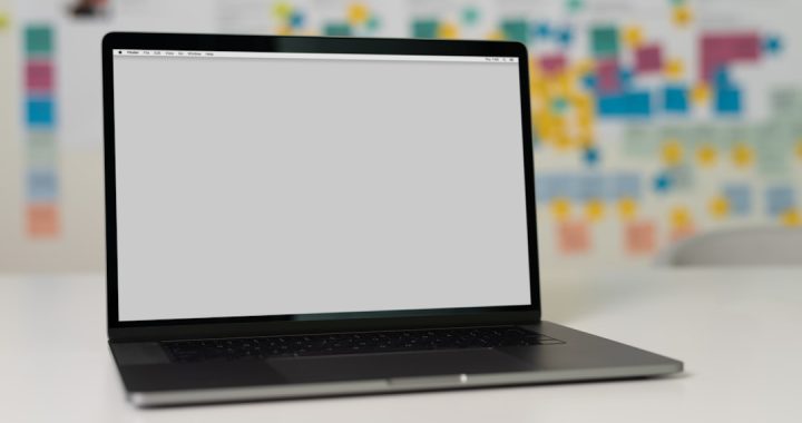 A laptop on a desk with an empty screen, background features sticky notes and papers organized on a wall.